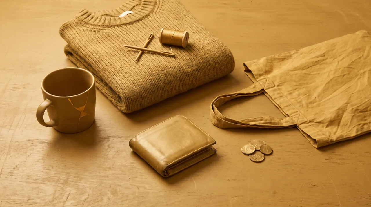 Bodegón en tonos dorados: mesa casi vacía con una taza de cerámica reparada, un jersey doblado con aguja e hilo y una cartera de piel gastada, metáfora del consumo consciente